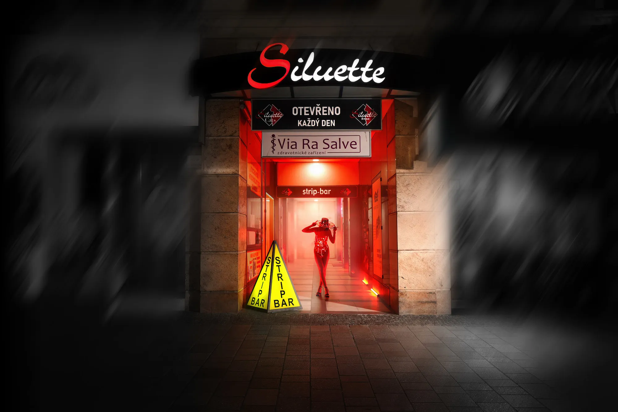 Noční pohled na vstup do pasáže v Brně s logem Siluette  nad průchodem. V této pasáži se nachází vstup do klubu Siluette. Vnitřek je osvětlen červeným světlem a hlouběji je viditelný nápis strip-bar. Uvnitř průchodu stojí tanečnice.