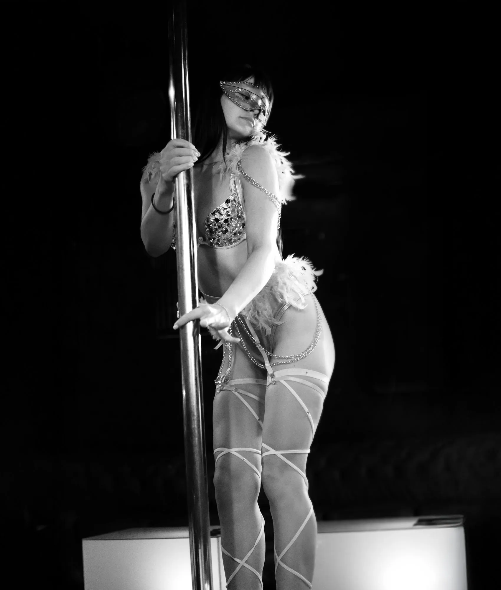 Tanečnice vystupující na pódiu u taneční tyče v interiéru klubu Siluette Brno. Má na sobě kostým s ozdobnými lesklými prvky, masku a péřové detaily. Fotografie je černobílá a zachycuje atmosféru živého večerního vystoupení.