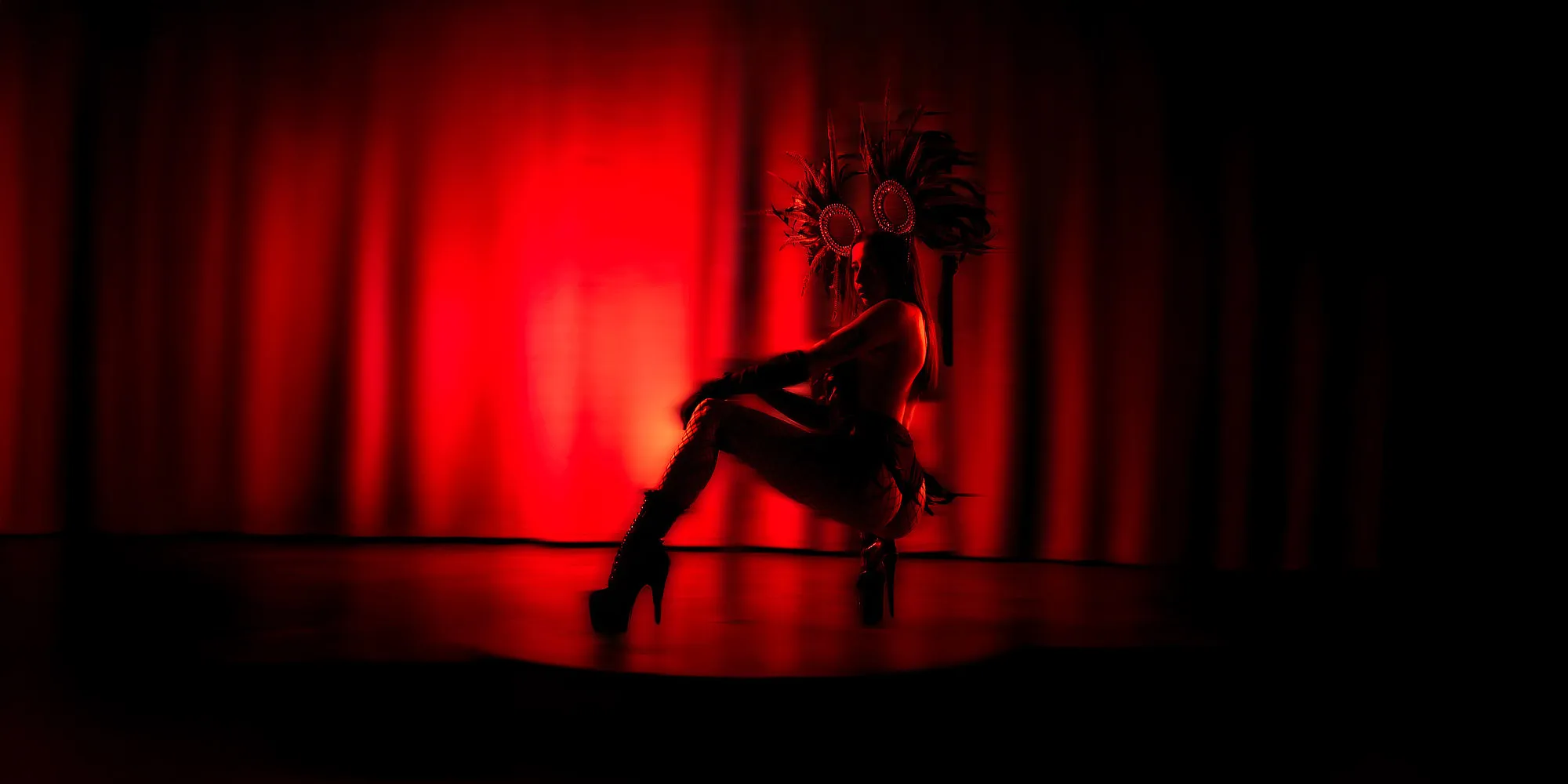 Tanečnice v výrazném šamanském kostýmu na pódiu před červeně nasvíceným pozadím a vytváří dramatickou siluetu plnou energie.